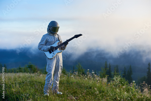Spaceman in white space suit and helmet holding white guitar standing on beautiful green mountain meadow in the morning, fog is rising up from the valley on background. Concept of music, astronautics.