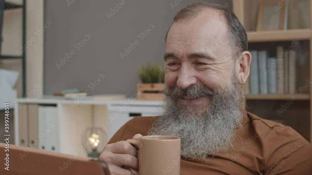 Close-up POV of kind Caucasian grandpa with grey beard holding mug, watching videos on tablet computer, then looking up and smiling on camera