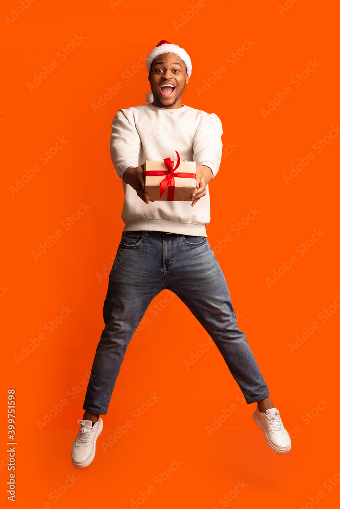 Gift For You. Excited African Guy In Santa Hat Jumping With Present Gift For You. Excited African Guy In Santa Hat Jumping With Present