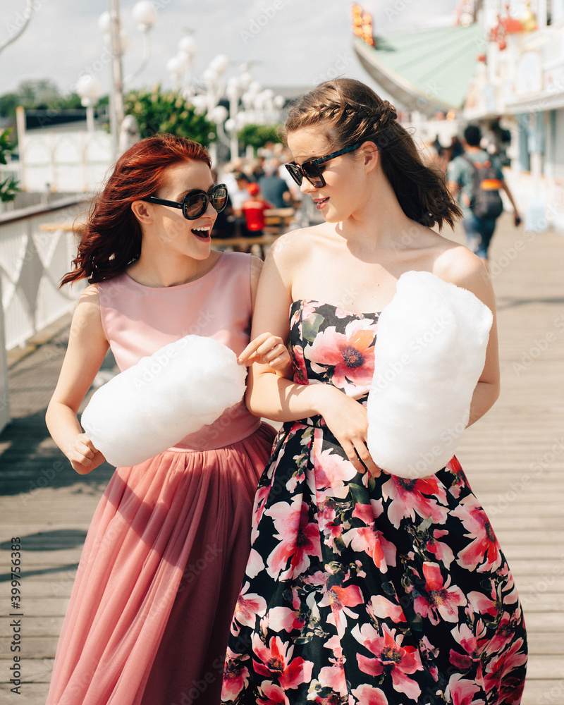 Naklejka premium Close up portrait of smiling excited girls with sunglasses and prom dresses holding cotton candy at amusement park. Teenagers in pink evening dresses on vacation. Birthday party idea.