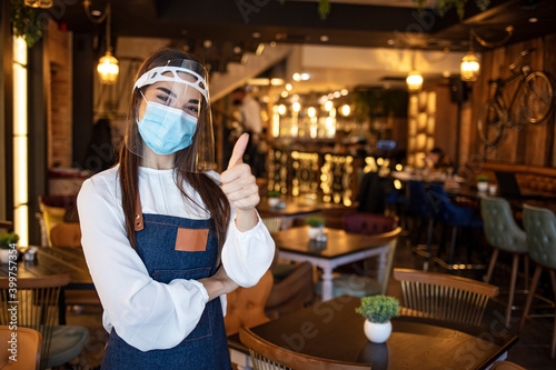 Portrait attractive waitress wear face mask and face shield showing OK sign with indoor restaurant background. New normal restaurant concept. Beautiful waitress working at a cafe