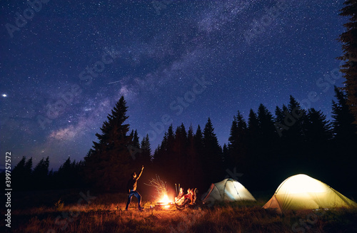 Man showing his friends Milky Way over tent city. Guys are sitting by the campfire on the background of the spruce forest and enjoying the evening starry sky. Outdoor recreation, touristm concept