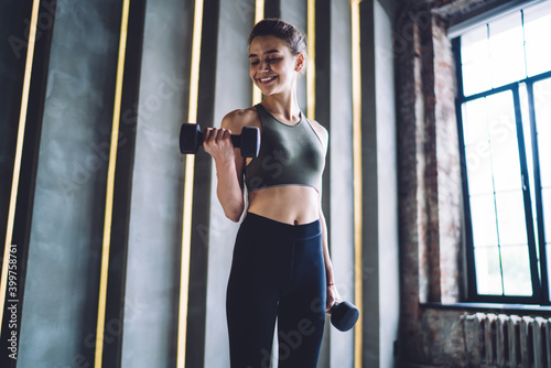 Happy caucasian female in active wear lifting dumbells and smiling enjoying reaching fitness goals, smiling woman spending time in gym using equipment satisfied with wellness and vitality