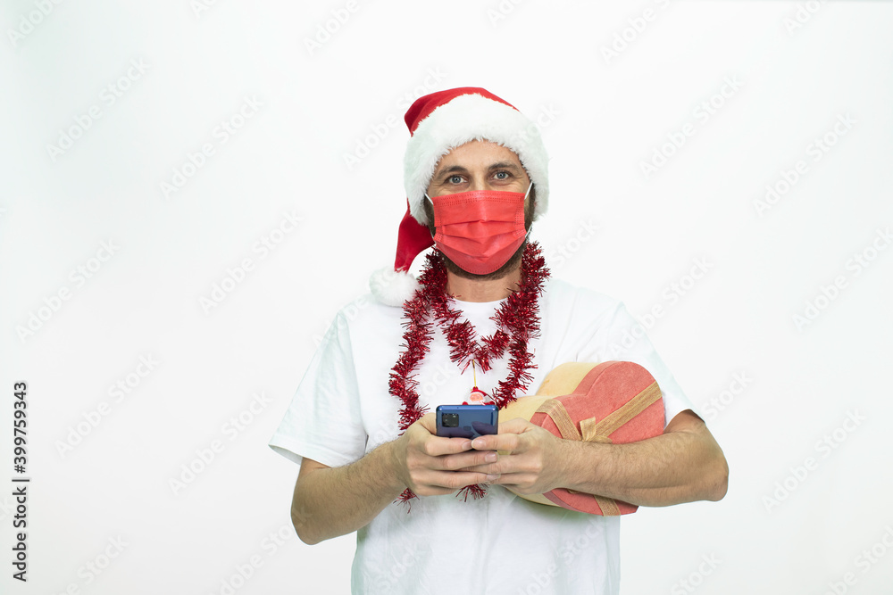 Man wearing a Santa hat. He has a red corona mask on his face. He is holding a gift box in the shape of a heart. Man wearing a Santa hat. He has a red corona mask on his face. He is holding a gift box in the shape of a heart.