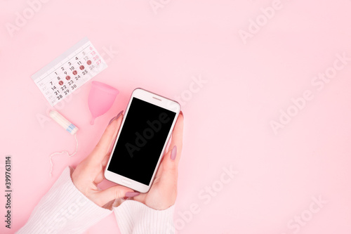 Phone on woman hands with menstrual calendar. Tampons and menstrual cup on pink pastel background. Concept of apps for critical days, menstruation. Top view,flat lay, copy space