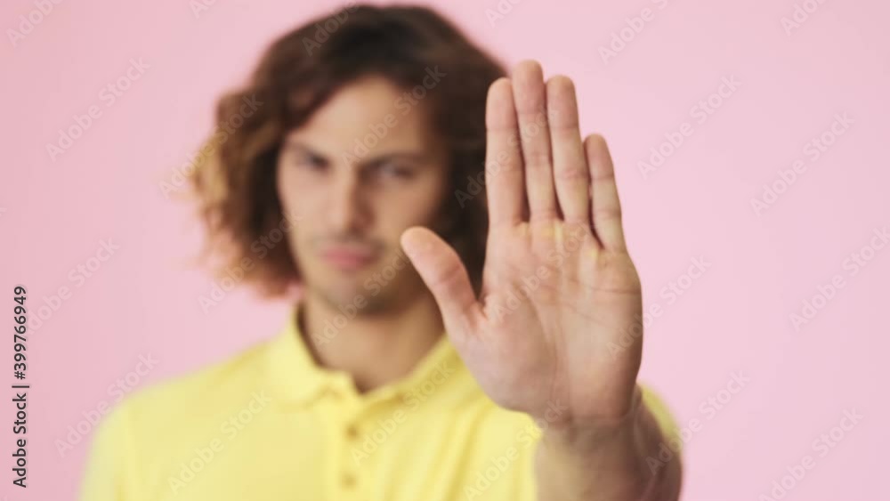 A close-up view of a serious curly young man is doing stop gesture showing his hand palm standing isolated over a pink background in the studio A close-up view of a serious curly young man is doing stop gesture showing his hand palm standing isolated over a pink background in the studio