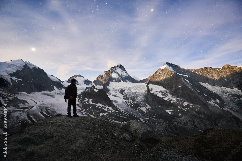 Unbelievably beautiful starry night illuminated by the Moon in Switzerland Alps, man standing and enjoying this peaceful scene of silent snowy peak Dent Blanche