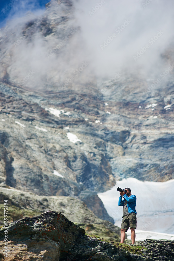 Male tourist using camera to take photo of beautiful mountains scenery on background. Mountain hiking, man reaching peak in sunny summer day. Concept of travelling and professional photography. Male tourist using camera to take photo of beautiful mountains scenery on background. Mountain hiking, man reaching peak in sunny summer day. Concept of travelling and professional photography.