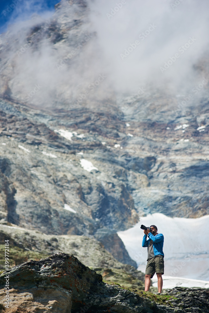 Male tourist using camera to take photo of beautiful mountains scenery on background. Mountain hiking, man reaching peak in sunny summer day. Concept of travelling and professional photography.