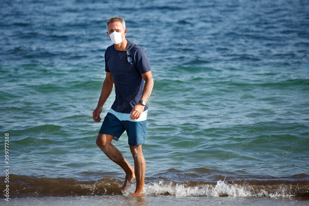 Uomo cinquantenne con mascherina protettiva e t-shirt blu sta in spiaggia e passeggia sereno di fronte al mare