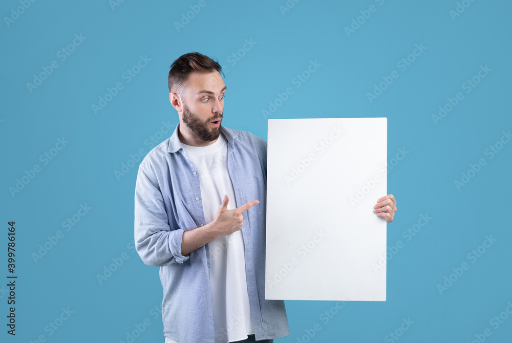 Surprised millennial man pointing at white blank poster on blue studio background, mockup for your ad design Surprised millennial man pointing at white blank poster on blue studio background, mockup for your ad design