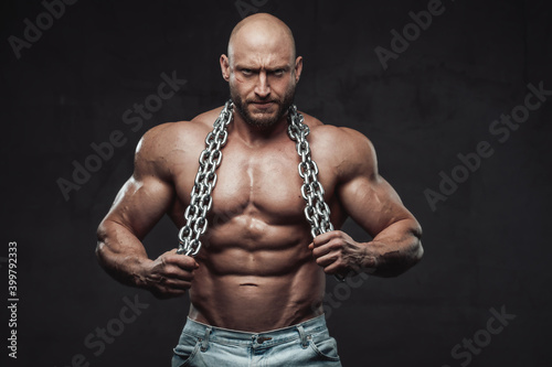 Tableau sur toile Handsome and proud man with muscular build and bald head poses with naked torso and steel chains in dark background looking at camera
