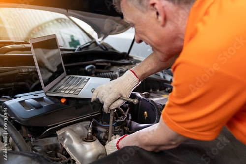 Professional male mechanic worker using laptop under hood of car at the repair garage. Male car mechanic working using wrench tool for examining, repair and maintenance at car service shop