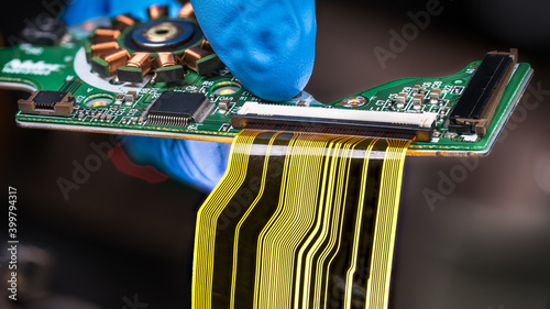 Flat plastic strip flexible cable on printed circuit board on a dark background. Hand in blue glove holding green PCB with integrated motor stator and electronic components as micro chip or resistors. photo