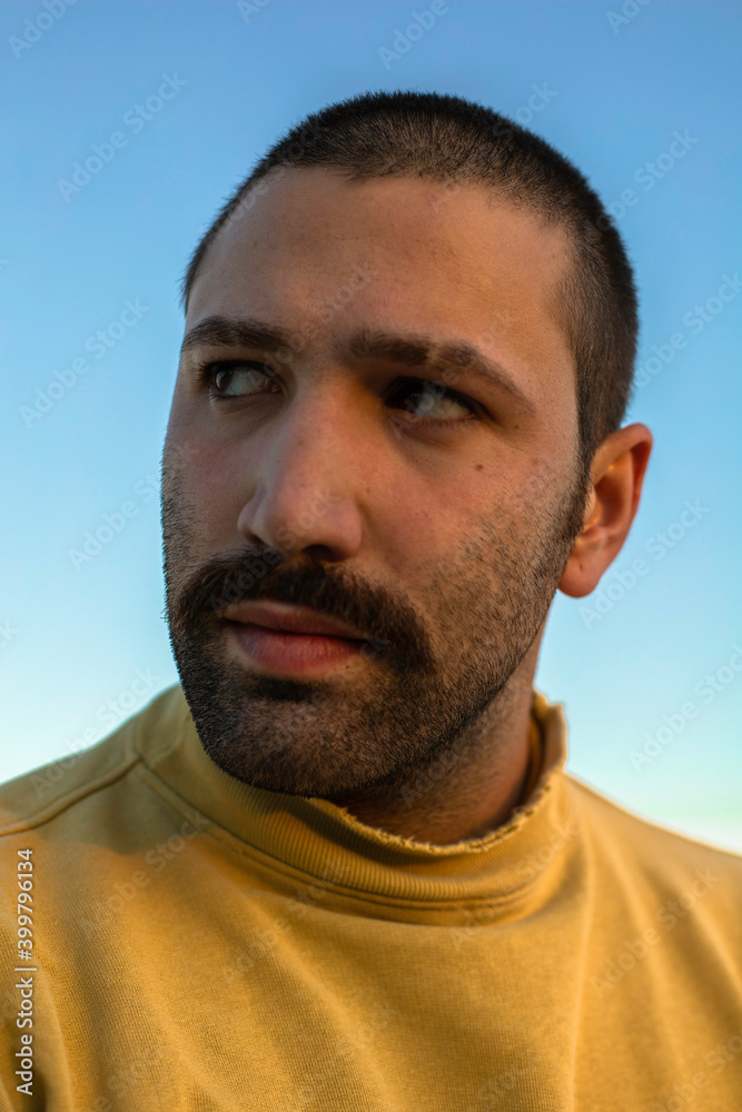 Fototapeta premium Portrait of young man with moustache looking away in front of blue sky wearing yellow jumper