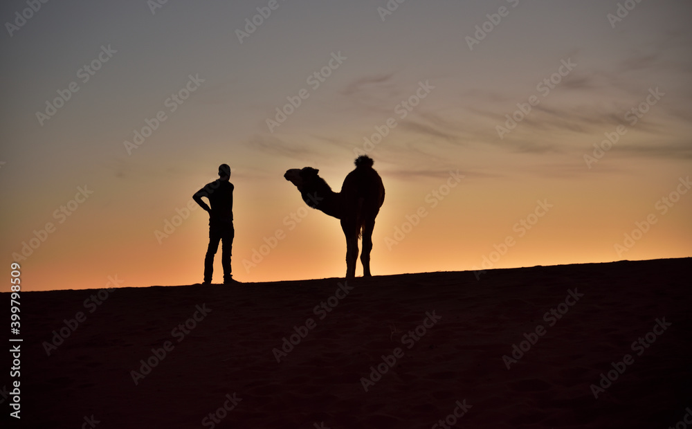 Silhouette einer Person und eines Kamels im Abendlicht auf einer Sanddüne in der Wüste