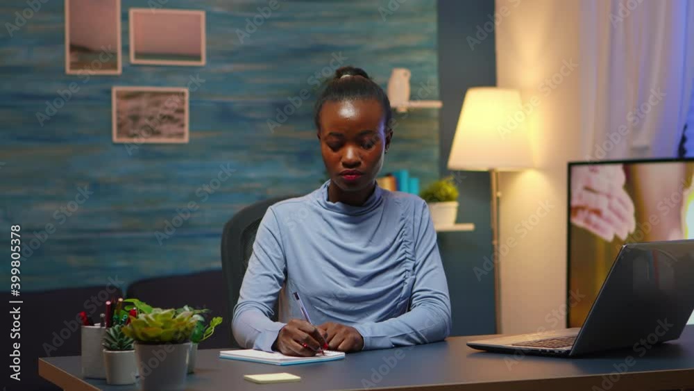 Black young business woman sitting at desk in living room workplace overtime and taking notes in notebook. African freelancer working from home late at night respecting deadline of project. Black young business woman sitting at desk in living room workplace overtime and taking notes in notebook. African freelancer working from home late at night respecting deadline of project.