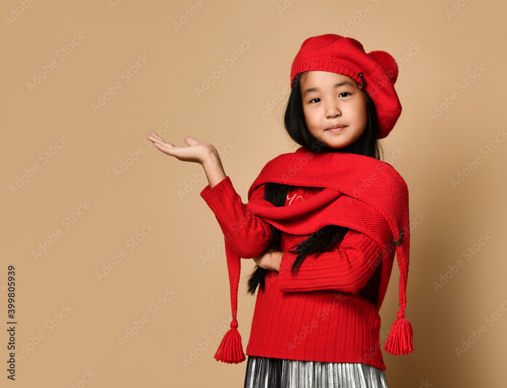 Side view of an Asian girl in a red hat, scarf and sweater, holding her palm and looking at the camera. Child posing on a beige background in the studio. Side view of an Asian girl in a red hat, scarf and sweater, holding her palm and looking at the camera. Child posing on a beige background in the studio.