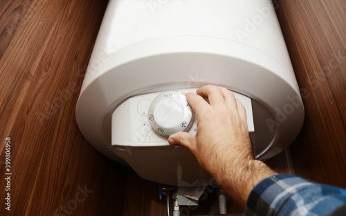 A man is switching on a domestic electric hot water boiler, water heater in a bathroom cabinet setting the temperature of the hot water for boiler efficient usage and energy saving.