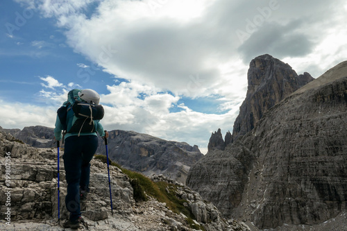 Woman with big backpack and sticks hiking in high Italian Dolomites. She goes up a steep slope. There are many sharp peaks in front of her. She is going up. There are a few trees around. Sunny day.