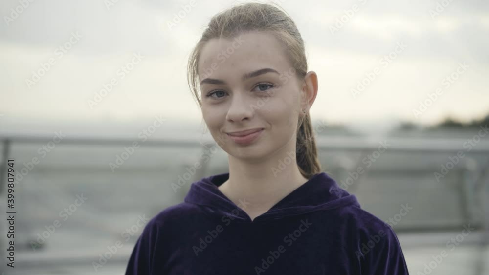 Portrait of positive Caucasian sportswoman posing outdoors on cloudy overcast day. Close-up face of smiling young woman looking at camera and winking. Lifestyle and beauty concept. Portrait of positive Caucasian sportswoman posing outdoors on cloudy overcast day. Close-up face of smiling young woman looking at camera and winking. Lifestyle and beauty concept.