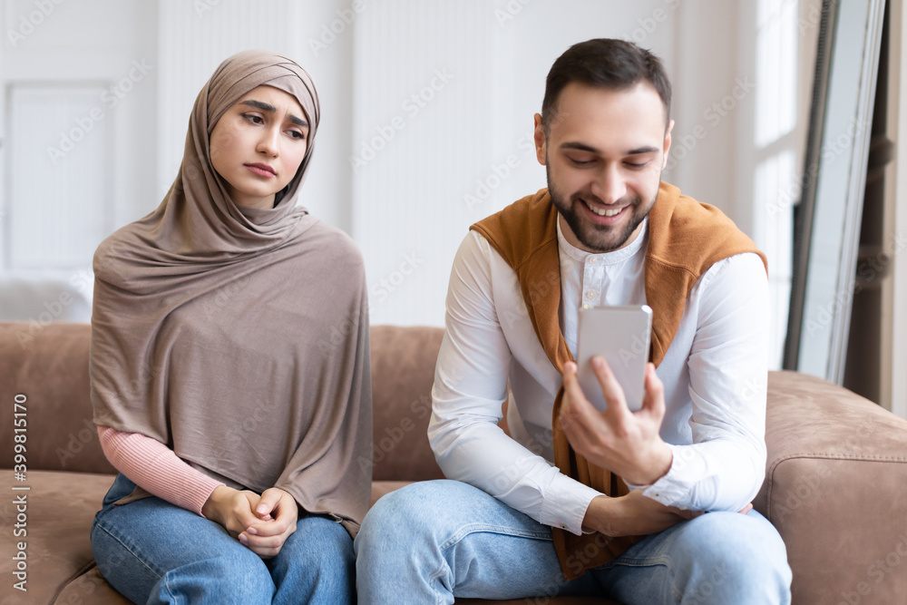 Unhappy Muslim Wife Looking At Husband While He Texting Indoor Unhappy Muslim Wife Looking At Husband While He Texting Indoor