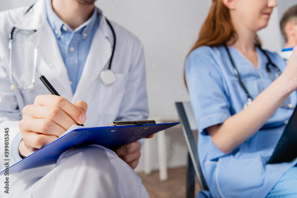 Cropped view of doctor with stethoscope writing on clipboard while sitting near nurse during meeting on blurred background Cropped view of doctor with stethoscope writing on clipboard while sitting near nurse during meeting on blurred background