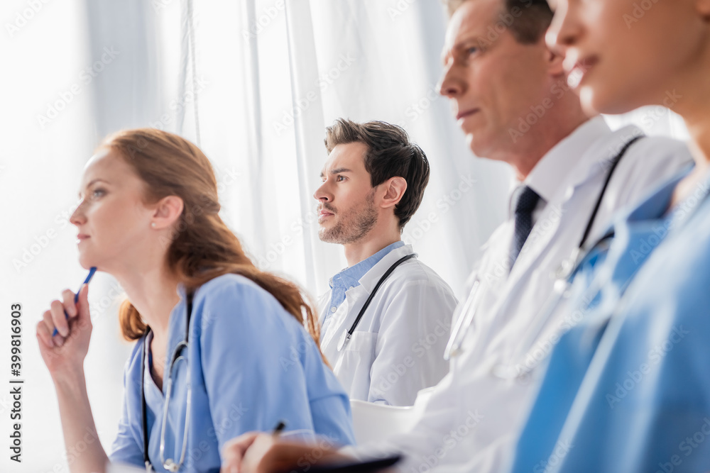 Redhead nurse with pen looking away near colleagues during meeting on blurred foreground