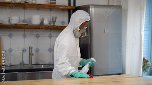 Foto Disinfector in protective suit and mask spraying detergent and wiping table in h