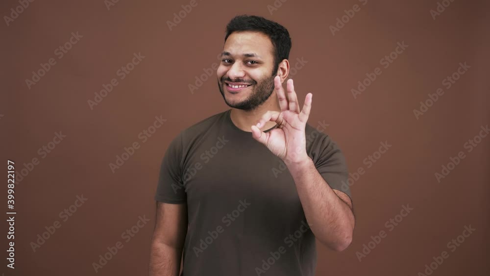 Awesome indian man gesturing Ok and nodding head, smiling to camera, brown studio background