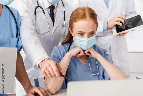 Fototapeta Naklejka Na Ścianę i Meble -  Redhead nurse in medical mask looking at laptop near multicultural colleagues standing behind on blurred foreground