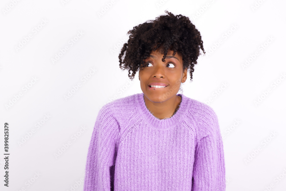 Amazed African American woman wearing purple knitted sweater against white wall bitting lip and looking tricky to empty space.