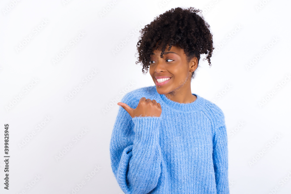 Charming oung beautiful African American woman wearing blue knitted sweater against white wall looking at copy space having advertisements Charming oung beautiful African American woman wearing blue knitted sweater against white wall looking at copy space having advertisements