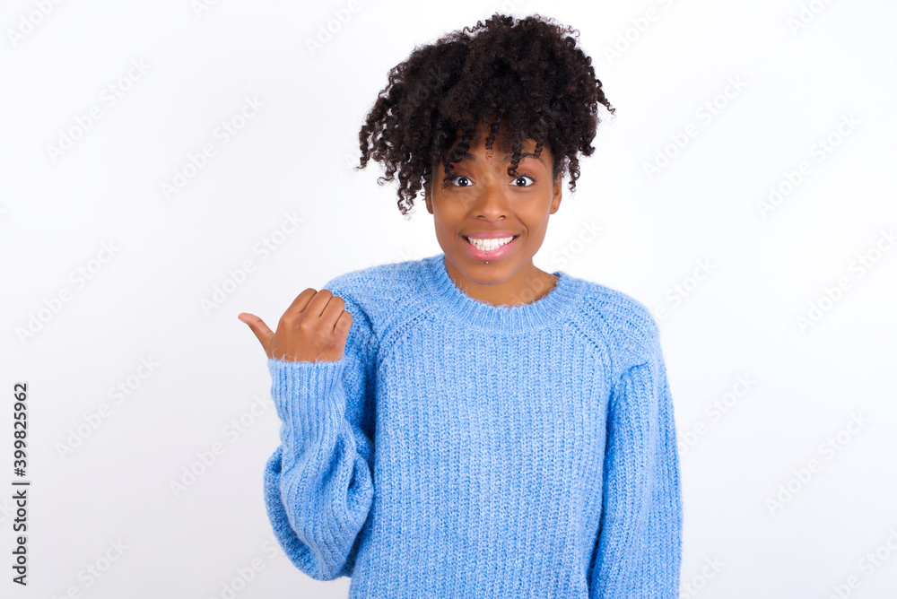 Lovely oung beautiful African American woman wearing blue knitted sweater against white wall pointing aside with forefinger, showing at copy space having news about bargains Lovely oung beautiful African American woman wearing blue knitted sweater against white wall pointing aside with forefinger, showing at copy space having news about bargains