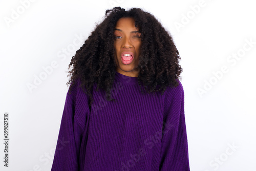 Young beautiful African American woman wearing knitted sweater against white wall,  sticking tongue out happy with funny expression. Emotion concept. © Roquillo