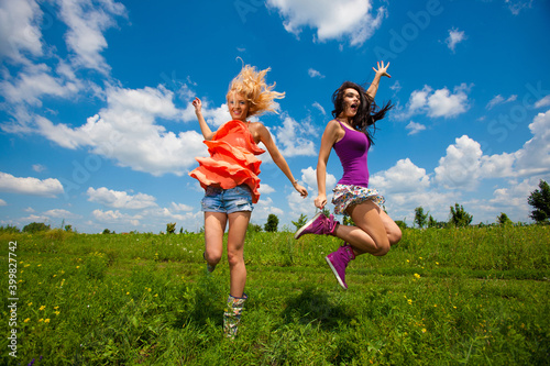 Two happy beautiful girls blonde and brunette twenty years old in colored dresses are jumping on a green field 