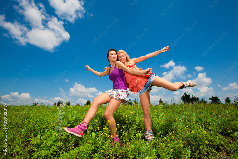 Two happy beautiful girls blonde and brunette twenty years old in colored dresses have fun on a green field with flowers Two happy beautiful girls blonde and brunette twenty years old in colored dresses have fun on a green field with flowers
