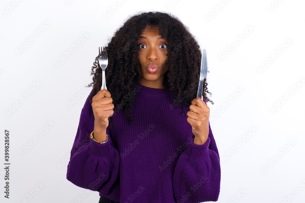 Model hungry Young beautiful African American woman wearing knitted sweater against white wall holding in hand fork knife want tasty yummy pizza pie Model hungry Young beautiful African American woman wearing knitted sweater against white wall holding in hand fork knife want tasty yummy pizza pie