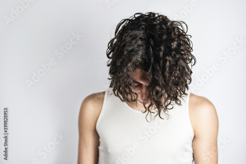 A teenage boy with long curly hair on a light background lowered his head, he seems to apologize for his misdeeds to his parents, asks to understand him and help to solve teenage problems