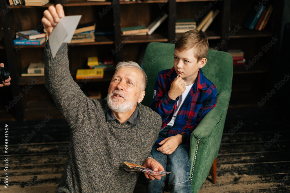Obraz premium Caucasian cheerful bearded gray-haired grandfather with his grandson enjoy memories watching family photo album sitting on armchair and floor at home in room with an authentic aristocratic interior.