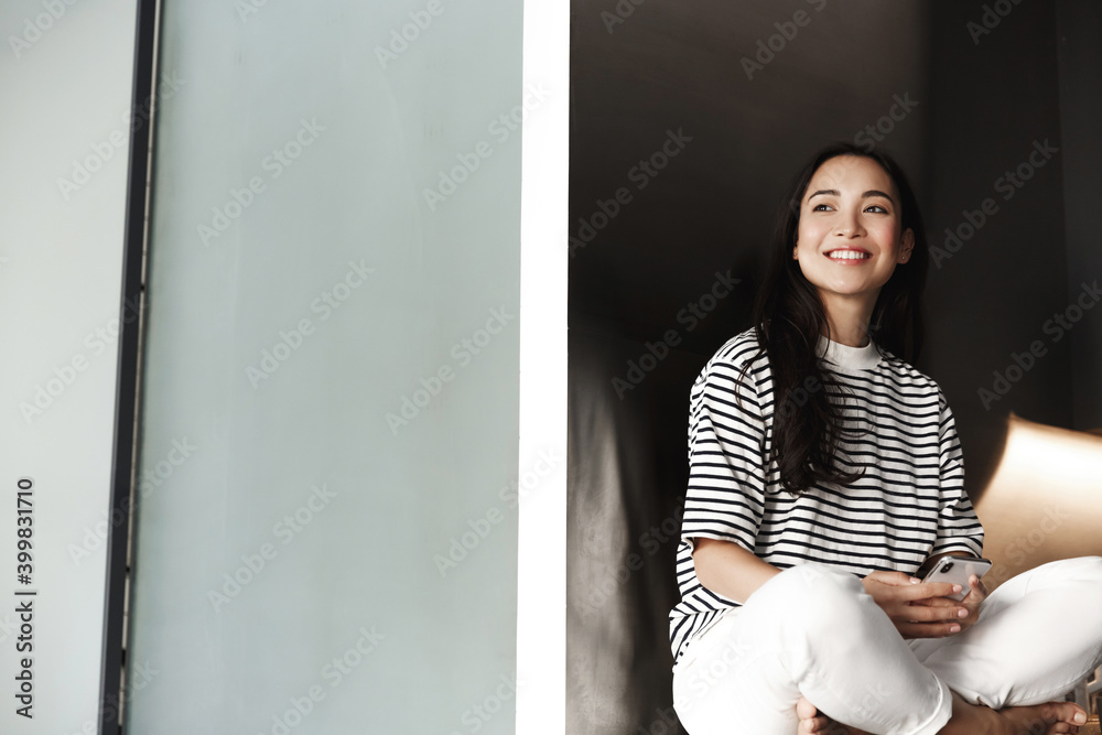 Asian woman rest at home with mobile phone. Girl sitting in her room and using smartphone, smiling and looking aside