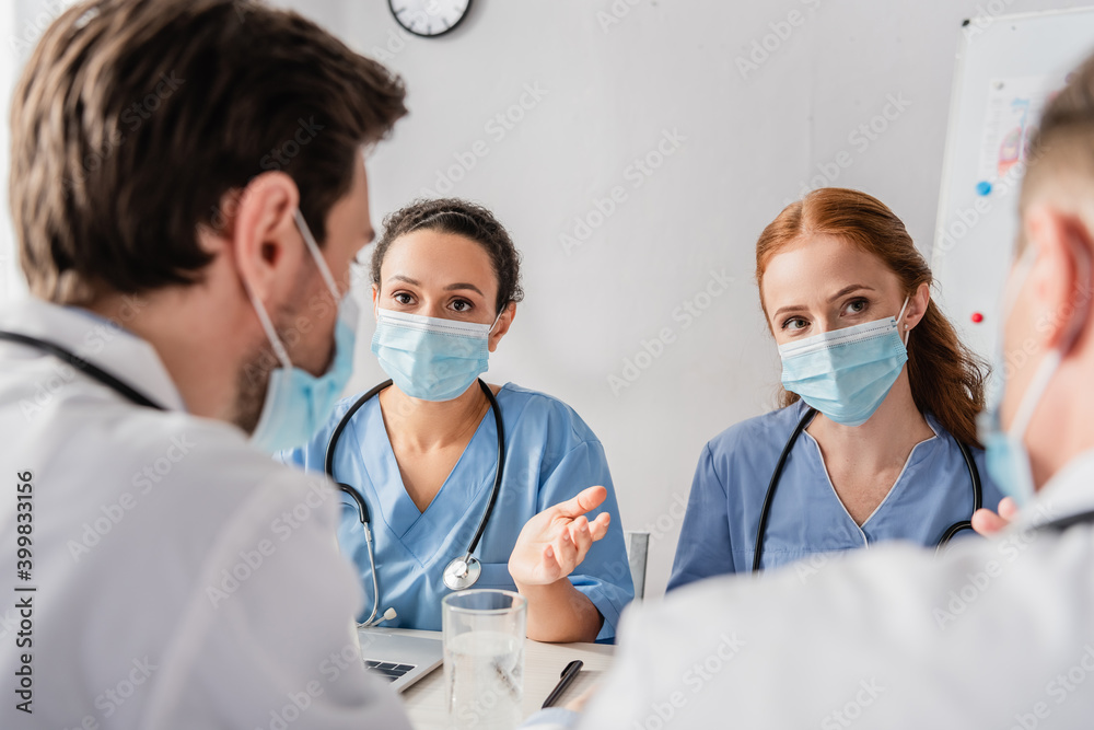 Multicultural nurses in medical masks looking at doctors while sitting at workplace on blurred foreground Multicultural nurses in medical masks looking at doctors while sitting at workplace on blurred foreground