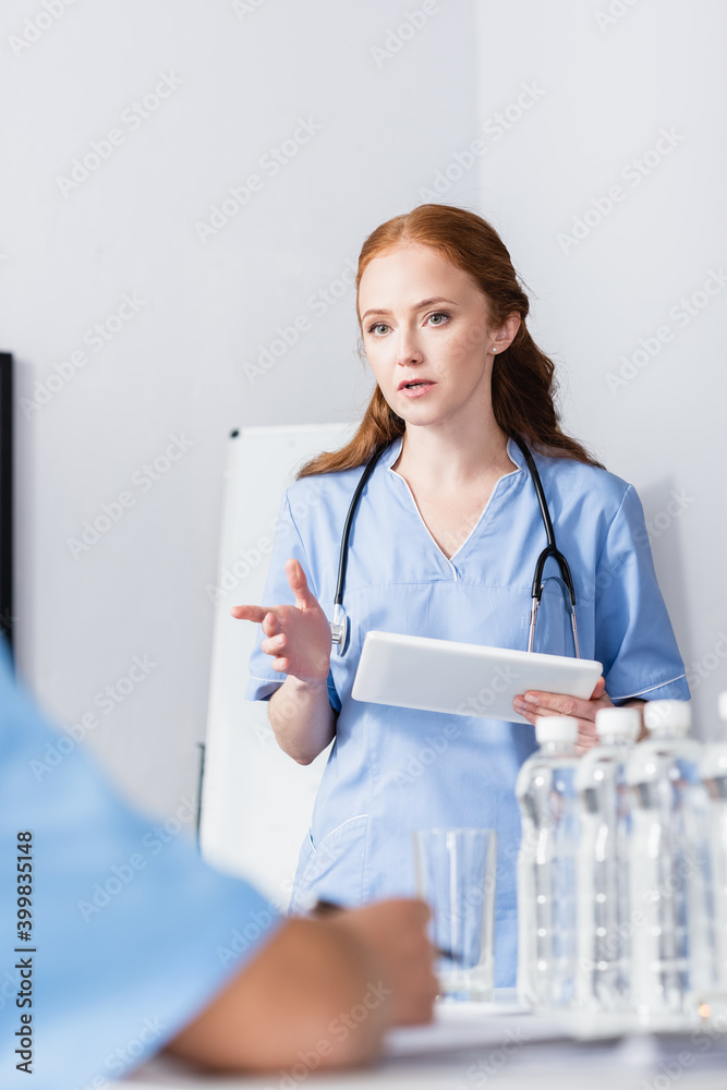 Nurse pointing with finger while holding digital tablet near colleagues and bottles of water on blurred foreground Nurse pointing with finger while holding digital tablet near colleagues and bottles of water on blurred foreground