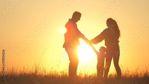 young mom  dad with a healthy daughter dance in a circle under warm sun  have fun in field. happy family  holding hands  plays in the park on grass  at sunset. Family and childhood concept.