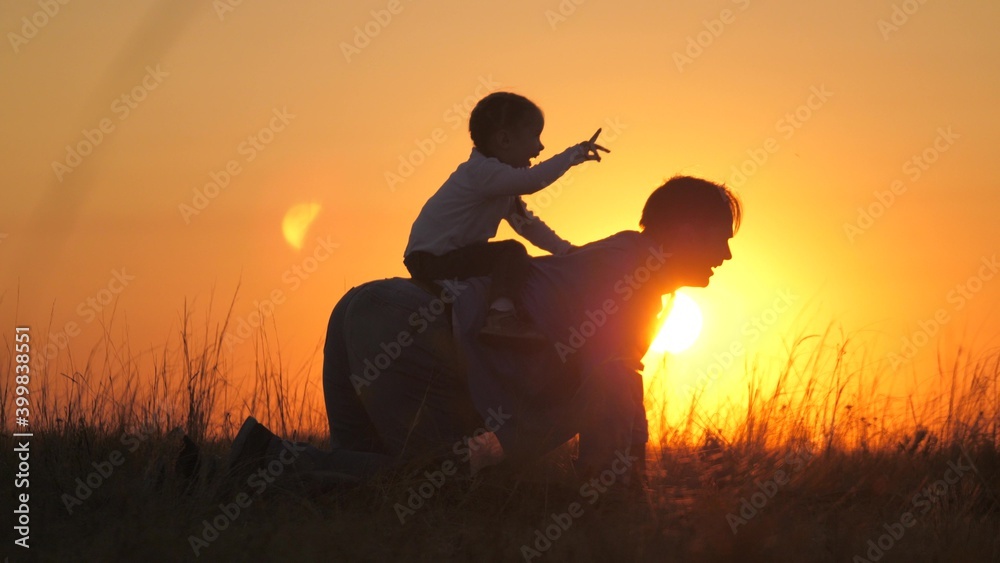 Dad plays with his little daughter in park on grass in rays of sunset. Father rides the baby on his back, he sits astride father, rejoices. Dad and little child are playing outdoors. Happy family