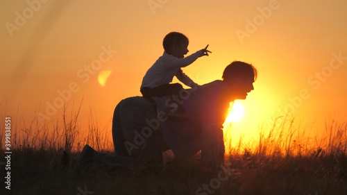 Dad plays with his little daughter in park on grass in rays of sunset. Father rides the baby on his back, he sits astride father, rejoices. Dad and little child are playing outdoors. Happy family © zoteva87