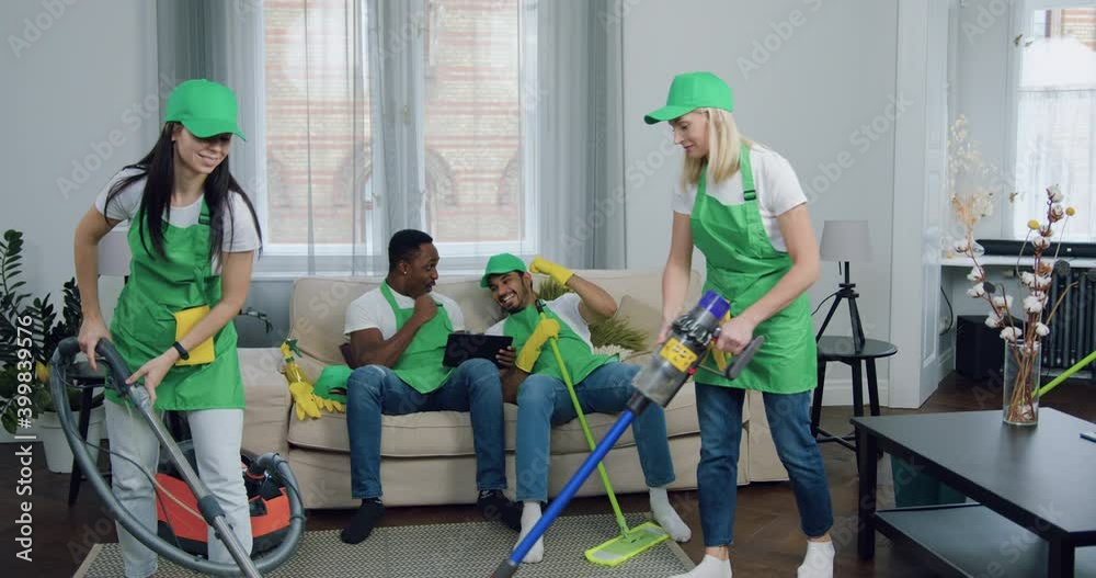 Good-looking happy cheerful multiethnic men-cleaners rejoicing from results of game on tablet pc while their hardworking skilled female partners cleaning carpet with vacuum cleaner in client's flat Good-looking happy cheerful multiethnic men-cleaners rejoicing from results of game on tablet pc while their hardworking skilled female partners cleaning carpet with vacuum cleaner in client's flat