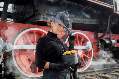 Fotografía Beautiful girl in a black dress and hat near an old steam locomotive and big iron wheels