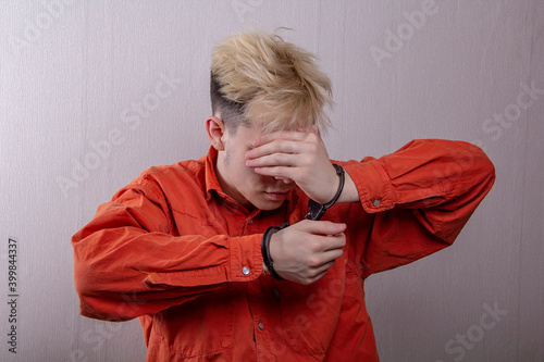Obraz na plátně Portrait of a teenager in handcuffs covering his face with his hands on a gray background, medium plan