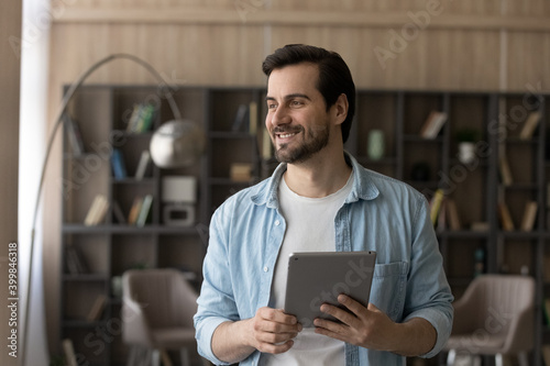 Happy millennial Caucasian man look in distance using modern tablet at home. Smiling young male user or client hold pad device, thinking or pondering. Online distant communication, technology concept.